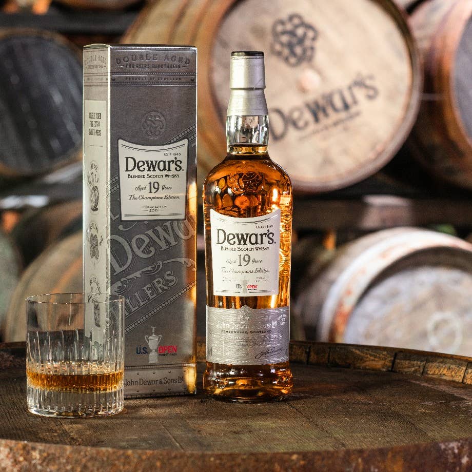 In front of the whiskey casks, a chest, bottle and glass filled with this 19-year-old Champion Dewar's Edition.